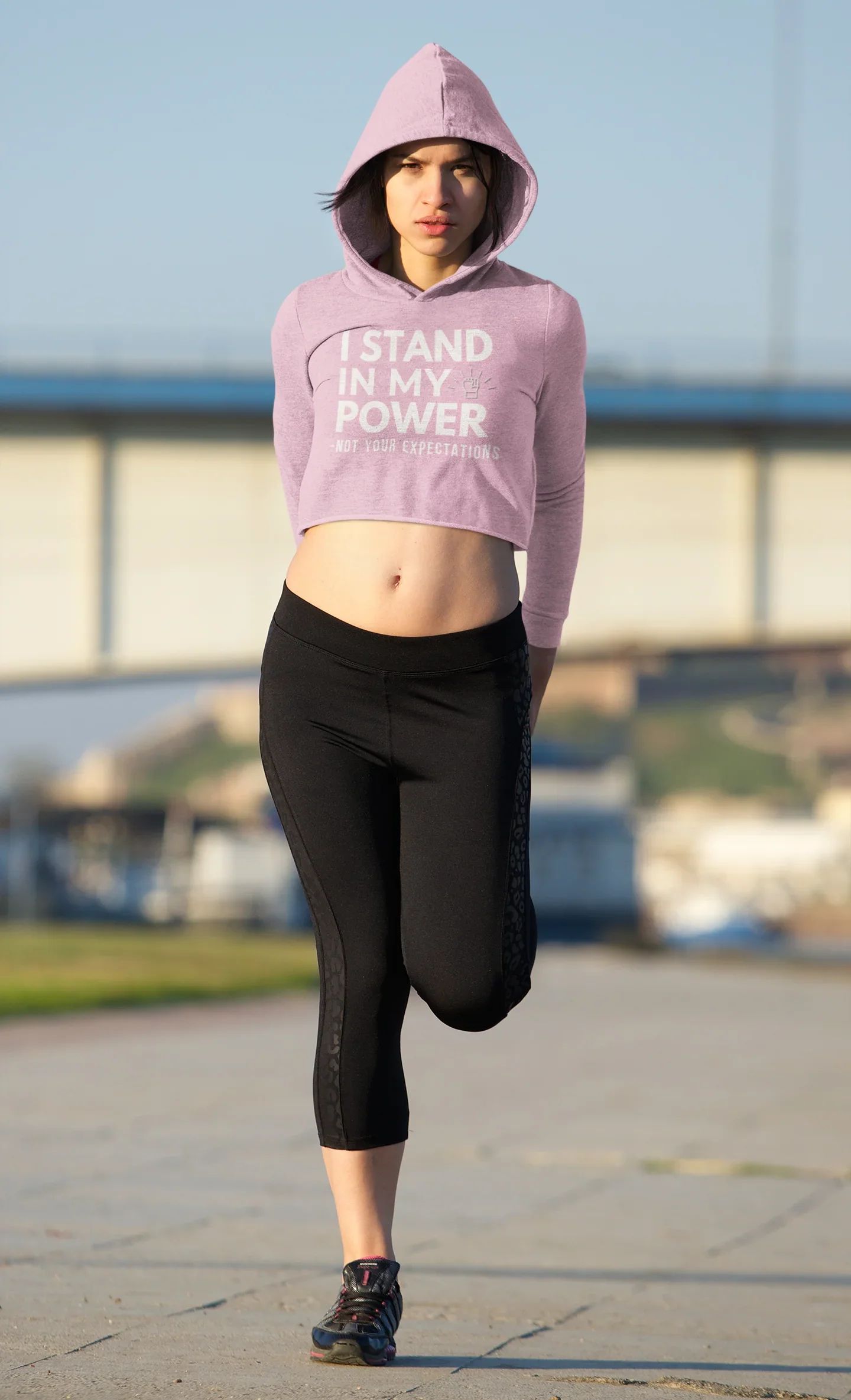 Woman in cropped purple hoodie with 'I stand in my power' text, stretching outdoors in activewear
